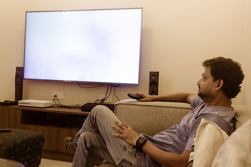 Omu chì vede a TV in casa in u salottu.L'omu asiaticu in maglietta blu pusatu nantu à un divano chì vede a televisione nantu à u divano.vista laterale.Oci, tecnulugia, mass media è cuncettu di persone